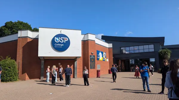 exterior image of the Seevic Campus. Clear blue skies, with the centre of the image being a brick hexagonal building, with the USP College logo on the front of it on a blue circle. to the right of the image is the main reception, and grey modern building with glass doors and glass extending to the ceiling with the logo printed on it. staff and students stood on the brick pathway outside Reception.