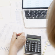 A female student working on a  laptop and holding a pen and making notes on a white paper.