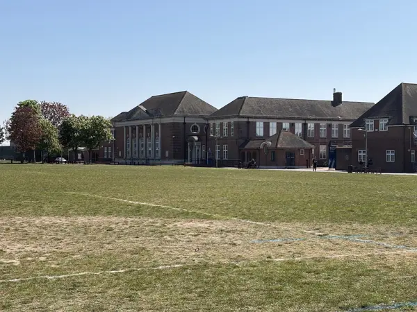 Exterior Image of the Palmer's Campus, showing a field in the foreground, with the main building in the centre of the image. a brick building, with the main building to the left of the image with pillars at the front of the building, and smaller brick building connected to it to the right of the image, and with grey gable roofs